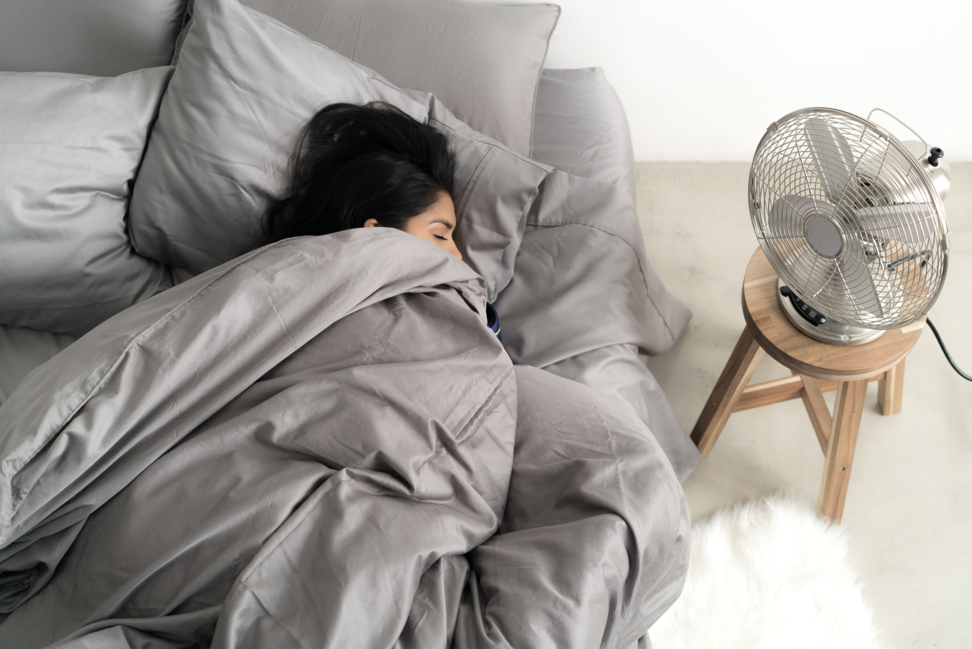 Woman sleeping comfortably under cool grey Miracle Sheets bedding beside a fan